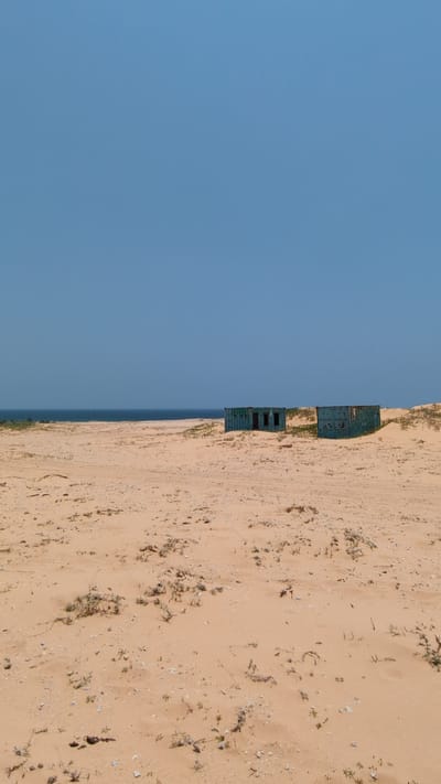 The image depicts a beach scene near Phuoc Dinh, Vietnam. On the right side of the frame, a sandy beach stretches out, marked with sparse vegetation and footprints. Two dilapidated, green metal structures, possibly shipping containers, are positioned on the edge of the beach, near the water. To the left is a clear blue sky. A sliver of the ocean is visible between the beach and the sky. The image is rotated ninety degrees counterclockwise.