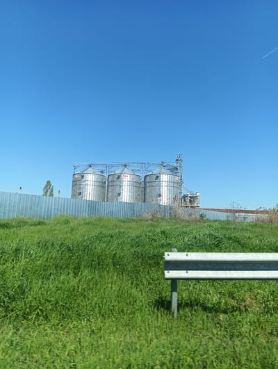 The image shows a landscape with grain silos in the distance, likely situated in an agricultural area. The location is Straldzha, Bulgaria. In the foreground is a field of tall green grass. A metallic guardrail is partially visible on the right side of the image. Behind the grass is a metallic fence, behind which stand three large, cylindrical, silver-colored grain silos with red markings. To the right of the silos are additional industrial structures. The sky is a clear, bright blue with a faint contrail from an aircraft visible at the upper-right corner of the image. The scene appears to have been captured from a moving vehicle.