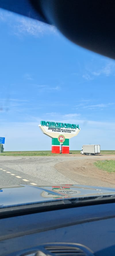 The image depicts a roadside scene in Камаево, Russia. A large sign with the words "TATARSTAN" in Cyrillic script and "WELCOME" in Russian and Tatar stands beside the road. The sign features the flag of Tatarstan. A small white truck is parked nearby. The view is from inside a vehicle, with the dashboard visible in the foreground. The sky is blue and clear. The ground is flat and grassy.
