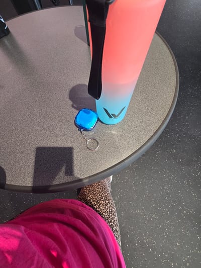 The image presents an indoor, high-angle view focused on a round table, objects on its surface, and the lower portion of a person. The table features a dark gray, speckled composite surface with a solid dark gray edge. Resting on the table is a tall, translucent water bottle with a color gradient transitioning from reddish-pink at the top to light blue at the base, partially filled with clear liquid. A black carrying strap is attached to the bottle, which also displays the brand name "WellMix." Next to the bottle is a small, translucent blue, faceted object, possibly a case or tracking device, connected to a silver keyring. Prominent shadows are cast on the table, including a distinct rectangular shadow (consistent with a mobile device) and shadows from the bottle and the blue object. The person in the foreground is dressed in bright fuchsia-colored fabric, which is wrinkled, and leopard-print leggings. A white shoe is partially visible on the left foot. The floor is dark gray with numerous light-colored speckles, indicative of a durable, possibly rubberized, surface found in commercial or public settings. The visual content of the image does not contain any identifying markers or contextual details to establish the specific location as Zirndorf, Germany.