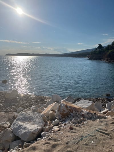 The image depicts a serene coastal scene in Potos, Greece, under a clear blue sky with the sun shining brightly. The foreground is dominated by a rocky and sandy shoreline, with large white and beige boulders scattered across the beach. The sea, a calm expanse of blue water, stretches out towards the horizon, reflecting the sun's dazzling rays, creating a shimmering effect on its surface. In the distance, a hilly coastline is visible, dotted with lush green trees and vegetation, suggesting a natural, undeveloped landscape. A few small boats can be seen floating on the water, indicating a peaceful, maritime environment. The overall impression is one of natural beauty and tranquility, characteristic of a seaside town like Potos.