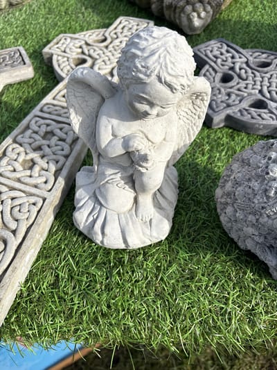 The image shows a close-up view of garden ornaments placed on artificial grass. The ornaments include a stone cherub lying on its side, a stone ball, and several Celtic crosses in various shapes and sizes. The setting appears to be outdoors, likely a garden or patio. The specific location is not evident in the image itself.