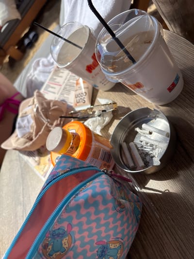 A close-up, overhead shot captures a collection of items scattered on a wooden table, likely outdoors given the natural lighting and shadows. In the foreground, a pink and blue patterned pencil case, featuring cartoon characters, is partially open. To the right of the pencil case is a silver ashtray filled with cigarette butts and ash. Behind the ashtray and pencil case are two white paper cups with clear plastic lids and black straws, suggesting beverages. To the left of the cups, a tan baseball cap is placed upside down. An orange plastic bottle with a white cap is visible between the cap and the pencil case. In the background, a white towel and parts of a lounge chair are visible, along with glimpses of what appears to be a menu. The overall impression is a casual, perhaps post-activity scene, consistent with a beach bar or resort setting. The items are arranged in a disordered manner on the wooden surface. Based on the provided information, this scene is located in Νέα Ηρακλείτσα, Greece.