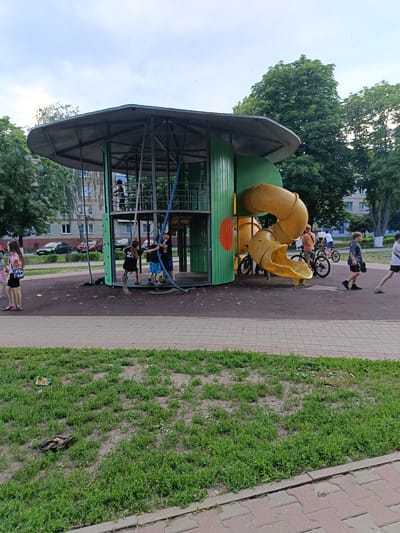 The image depicts a modern, colorful playground structure with a yellow spiral slide and green siding, located in an urban park. Several children are actively engaged with the playground equipment. One child is climbing a rope, while others appear to be inside or near the structure. In the background, trees and apartment buildings suggest a residential area. A few adults and children are visible in the distance, some with bicycles. The ground is a mix of soft play surfacing and paved walkways, with a strip of grass in the foreground containing some litter. The overall scene suggests a typical summer day at a public park in the city of Gubkin, Russia.
