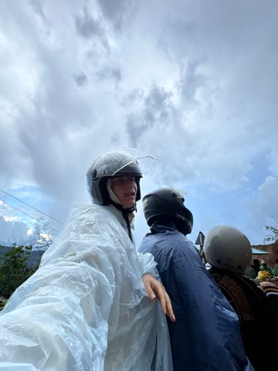 The image shows three people on a motorcycle in Nao Sẻ, Vietnam, wearing raincoats and helmets as it is raining. The person in the foreground is sticking their tongue out playfully, with water droplets on their clear helmet visor. The person in the middle is wearing a dark blue raincoat and a black helmet. The third person, mostly obscured, is wearing a light grey helmet. The background shows a cloudy sky and some greenery. The overall atmosphere is casual and adventurous, despite the rainy weather.