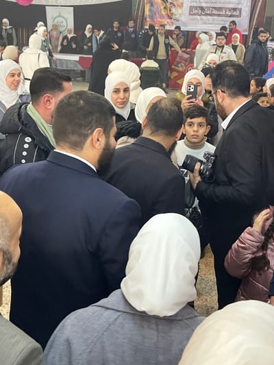 This image captures a crowded indoor event in Kafr Batna, Syria. Approximately 30-40 individuals are visible, predominantly adults. In the foreground, the backs of three individuals are prominent: a man in a dark navy suit on the left, another man in a dark suit to his right, and a woman in a grey coat with a white hijab on the far right.
In the mid-ground, a young boy with short dark hair, wearing a light beige turtleneck sweater, looks intently towards the left. To his right, a man in a dark suit holds a black DSLR camera with a substantial lens, pointing it towards the boy or the central activity. Another woman, wearing a white hijab, is positioned behind the boy, holding a smartphone with a light-colored case up as if recording. Numerous other people, including women wearing hijabs and men with beards, are present throughout the scene, observing the activity.
The background features a large white banner stretched across a wall, displaying Arabic text that translates to "An event of Safety and Hope." A smaller banner with green text is partially visible on the far left. Beneath the banners, several individuals stand behind tables covered with red and white tablecloths, indicating display or information booths. The flooring appears to be a light-colored tiled surface. The overall environment suggests a community gathering or organized public event.