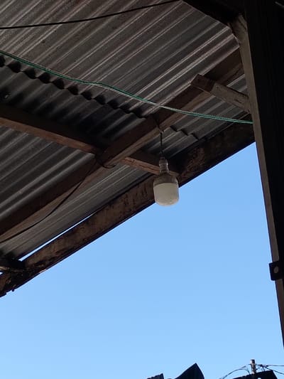 A corrugated metal roof covers the upper frame, supported by weathered wooden beams. A single, frosted light bulb hangs suspended from the wooden structure. An electric wire runs along the underside of the roof. In the background, a clear blue sky provides a contrasting backdrop. A portion of a dark support beam is visible on the right edge of the frame. A glimpse of other structures is seen below.