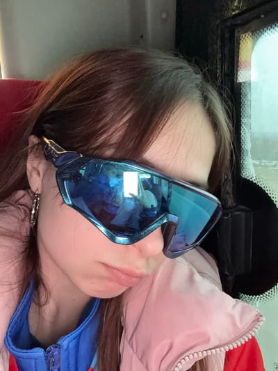 A young woman is lying down, possibly sleeping, with blue sunglasses covering her eyes. She appears to be in a vehicle, based on the window and vehicle interior visible. She is wearing a blue jacket with a zipper and a pink jacket is also visible. The image was taken in Челва, Russia.