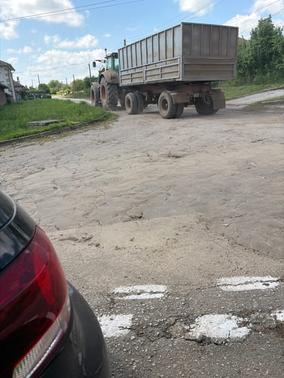 The image shows a tractor with a trailer driving on a cracked asphalt road in Kozlovets, Bulgaria. A car with a red taillight is partially visible in the lower right corner of the frame. The road has partially obscured white lane markings. Vegetation borders the road. Buildings and trees are visible in the background. The sky is visible with clouds.