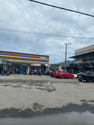 The image depicts a street scene in Lampisang, Indonesia, under an overcast sky. Prominently featured is an Indomaret convenience store, its facade marked by vertical stripes of blue, yellow, and red. The store appears to be open, with visible merchandise and people inside. Parked motorcycles and cars line the street in front of the store, with a red hatchback and a black SUV being easily identifiable. The street surface is concrete, showing signs of dampness or recent rain. Overhead, power lines stretch across the sky. In the background, a building and a mountain are visible.