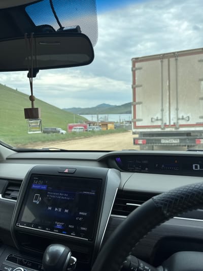 The image shows the interior of a car looking out at a road and a body of water. A large truck is visible ahead, and the road appears to be unpaved. In the background, there are hills and a cloudy sky. The car's dashboard is visible, with a navigation screen displaying "Bluetooth Audio" and song information. The time is shown as 14:01. The car is likely in or near Khuzhir, Russia, given the presence of Cyrillic text on the screen.