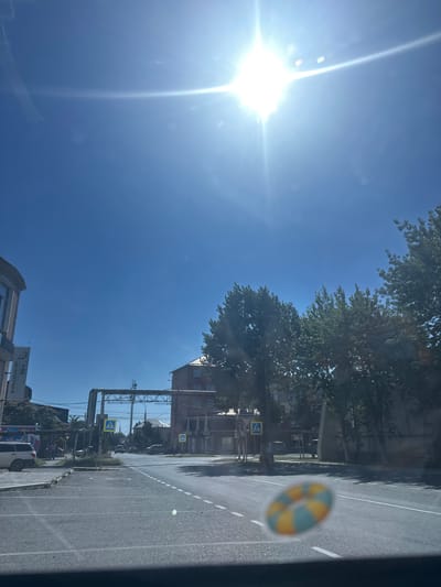 The image captures a sun-drenched street scene in Temryuk, Russia. A bright sun with lens flare dominates the upper portion of the frame, casting a strong light. The foreground shows a road with clear lane markings and a pedestrian crossing. A small, circular, blurred sticker with a yellow and blue checkered pattern is visible on the lower right of the windshield.
On the left side of the street, a building with an arched window and a vertical sign reading "TOP Автозапчасти" (TOP Auto Parts) is partially visible. Further down the street, several cars are parked and driving. In the background, trees line the street, interspersed with buildings, including a red brick structure with multiple windows. Two yellow pedestrian crossing signs with a blue border are prominently displayed at intersections. Overhead, a large metal structure, possibly part of a utility or infrastructure system, spans across the road. People are seen walking on the sidewalk in the middle ground. The overall environment suggests a typical day in a town or small city with moderate traffic and pedestrian activity.