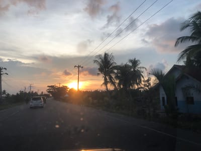 The media file depicts a rural road in Keudebagok Dua, Indonesia, during a sunset. The sun is low in the sky, casting a warm, orange glow and creating lens flare on the camera lens. The sky is a mix of soft grey and orange clouds, with power lines crisscrossing above. On the side of the road, there are scattered houses, palm trees, and other vegetation. A white car is driving away from the viewer on the road, with another car behind it. A person is walking on the left side of the road. The scene evokes a sense of tranquility and the end of a day. The overall atmosphere is peaceful, with the natural beauty of the sunset dominating the visual.