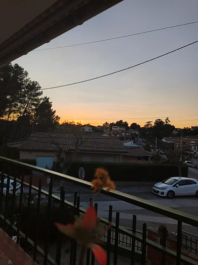The image shows an evening or sunset view from a balcony in Sant Esteve Sesrovires, Spain. The sky is transitioning from a blue to orange hue. In the foreground, a dark metal railing borders the balcony. Part of a blurred plant with red and orange tones is visible. The middle ground shows a street with a white car parked on the right and a hedge running along the houses. The houses have tiled roofs. Further back, the skyline is punctuated by trees and rooftops, with the setting sun partially obscured by the landscape. An overhead wire stretches across the sky.