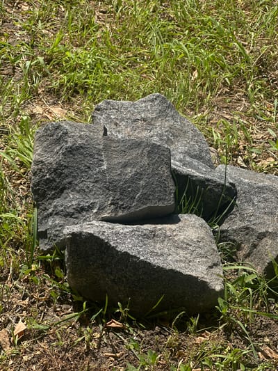 The image displays a close-up, overhead view of several large, rough-textured grey rocks resting on a bed of green grass and dry soil. The rocks are irregularly shaped and appear to be igneous, possibly granite, with visible flecks of lighter minerals. Some of the rocks are positioned to create small crevices where blades of grass grow. Sunlight casts shadows across the rocks and the surrounding ground, indicating it is daytime. The setting appears to be outdoors, in a natural environment, consistent with the provided location context of Yablunivka, Ukraine. There is no discernible activity or event taking place; the image focuses on the static arrangement of rocks and vegetation. No text is visible in the image.