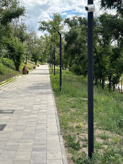 The image shows a paved walkway in a park-like setting, likely in Otradnoye, Russia. The walkway is constructed of light gray rectangular pavers. To the right of the walkway, there is a grassy area with some yellow wildflowers and several black lampposts. One lamppost in the foreground has a white security camera mounted near its top. Further down the walkway, a person in a red and white shirt is seated on a wooden bench. The park has a gentle incline to the left, with trees and green foliage. The sky above is partly cloudy.