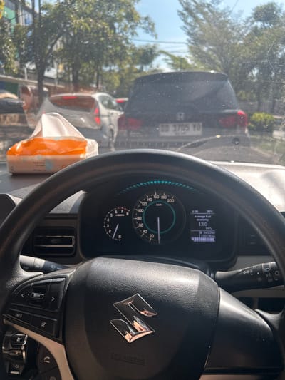 The image is taken from inside a vehicle, looking forward through the windshield. The dashboard and steering wheel are prominent in the foreground. A box of Guardian facial tissues is visible on the dashboard. The vehicle is in traffic, with several other cars ahead. The license plate of the car in front reads "DD 1775 YO". Trees and buildings are visible outside the car, suggesting an urban environment. The sky is clear and bright. The dashboard displays include a speedometer, tachometer, and a digital screen showing "Average fuel economy 13.0 km/L", "643.0 km", and "120136 km". The time is displayed as "6:36" and the temperature as "39°C". The overall scene depicts a moment of waiting in traffic, likely during the daytime. The location is identified as Pandang-Pandang, Indonesia.