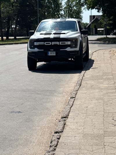 A white Ford F-150 Raptor pickup truck with black racing stripes on the hood and roof is parked on the right side of a paved road in Rudamina, Lithuania. The truck is facing forward and its front grille displays "FORD" in large block letters. A license plate reading "LSB 922" is visible on the front bumper. The truck is positioned next to a sidewalk made of textured concrete slabs. The road is asphalt and appears to have some minor cracks and debris. In the background, there are trees with green foliage and a building with a dark roof and windows. The sky is clear and blue. The lighting suggests it is daytime, with sunlight casting shadows.