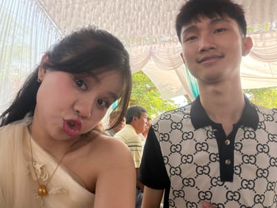 Two young adults pose cheek to cheek under a white tent-like structure, presumably at a wedding venue. The male, on the left, wears a black and white patterned shirt and smiles at the camera. The female, on the right, puckers her lips and wears a cream-colored dress with a gold necklace. An older man in a pale striped shirt stands in the background. The location is likely the city of Bình An, Vietnam.