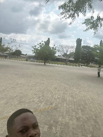 The scene depicts a large, open paved area under a partly cloudy sky in Ozuaha, Nigeria. The ground is covered with light-grey interlocking pavers, across which a faded yellow line extends from the lower-left. The camera operator's head is partially visible in the lower-left foreground. In the mid-ground, a black and white striped curb delineates the paved area from a grassy section containing numerous trees. These trees include tall, columnar specimens, a tree with sparse light-colored foliage, and several with dense green canopies. Behind the trees, multiple low-rise buildings with light-colored walls and dark roofs are visible. On the far right, a lone individual wearing a light-colored long-sleeved shirt and dark trousers stands on the paved surface, facing away from the camera. Tree branches extend into the top of the frame.