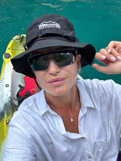 A woman is kayaking on turquoise water, likely in the Bay of Kotor near Bezuje, Montenegro. She wears a black bucket hat with an "OUTDOIRS" logo, large sunglasses, and a white linen shirt. A yellow kayak with a red life vest is visible beside her. Her hand, with light pink nails, adjusts her hat. The water reflects the surrounding cliffs.