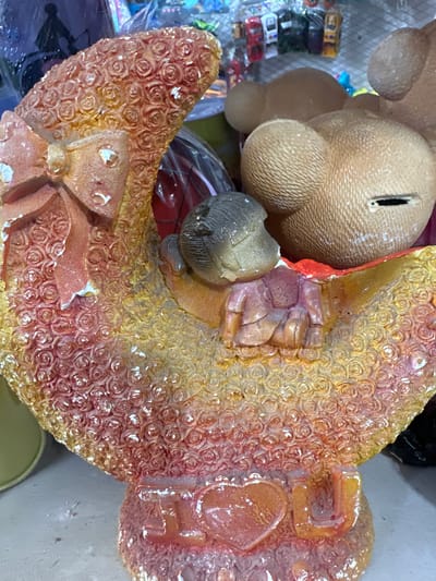This is a close-up, eye-level, medium shot of a decorative item. The item appears to be a figurine of a crescent moon adorned with rose patterns and a bow on the left side. A small figurine of a child is nestled within the crescent moon. Behind the moon is a portion of what appears to be a large, light brown, bear-shaped object with a coin slot, suggesting it might be a piggy bank. The background is out of focus and shows a cluttered collection of items, possibly in a shop or market setting. The item is positioned on a light-colored surface, which could be a shelf or table. The overall impression is one of a whimsical, decorative piece. There is no indication of the location being in Tokmok City, Kyrgyzstan.