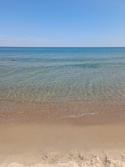 The image depicts a tranquil scene at a beach in Zatoka, Ukraine. The horizon is clearly defined, separating the vast expanse of a clear, bright blue sky from the deep blue of the sea. The water's surface is gently rippled, with sunlight creating shimmering patterns on the shallow seabed, which is covered in small pebbles and shells. The shore is composed of light brown sand, with a small wave lapping at the edge of the water. The overall impression is one of peace and natural beauty, characteristic of a sunny day at the seaside.