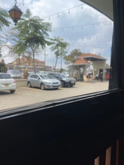 The scene depicts an outdoor environment in Gatongora ward, Kenya, viewed through a dark window frame that obscures the lower portion of the image. A paved area, likely a parking lot or plaza, occupies the midground. Several vehicles are parked: a white car (rear visible, left), a silver hatchback (center), and two dark sedans (center-right). To the right, two structures with reddish-brown, multi-tiered tiled roofs are present; one is an enclosed building, possibly a kiosk, and the other an open-sided shelter. Undefined human figures are faintly discernible near these structures, with one light-colored chair visible outside the shelter. Green-leafed trees are distributed across the midground and background. A string of decorative lights spans horizontally across the upper midground. An amber-colored, antique-style lantern hangs in the upper-left quadrant, partially obscured by foliage. Multi-story buildings form the distant background under an overcast sky. No specific actions or interactions are clearly identifiable due to image blur and distance; the scene suggests a public or commercial area with parked vehicles and people present.