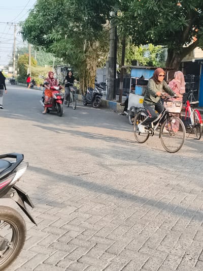In Barengkrajan, Indonesia, during daylight, a street scene unfolds with several individuals and vehicles. On the right side of the paved street, a woman wearing a brown hijab and sunglasses cycles on a bicycle with a white basket. She is dressed in a dark green long-sleeved shirt and dark pants. Behind her, another woman wearing a pink hijab and a floral patterned dress is also on a bicycle, which is partially obscured. To the left of these cyclists, a red scooter is occupied by a woman in a red and orange patterned hijab and dress. She is looking forward. Further to the left, a person wearing a black hijab and gray pants walks away from the camera. Various parked motorcycles are visible on the side of the road. Trees and foliage line the street, with power lines and poles also present. Buildings are visible in the background. The ground is paved with interlocking brick pavers.