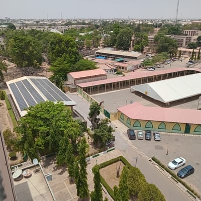 The image is an aerial shot of a location in Jama A, Nigeria, and features a mix of buildings, trees, and vehicles. The cityscape is visible in the background, with a skyline punctuated by various structures.
The foreground is dominated by buildings, with a structure covered in solar panels on the left side of the image. Next to it is a cluster of trees. There are more buildings with red roofs and tan-colored exteriors, some with a long open corridor. One of the buildings has a large white tent erected next to it.
A parking area is visible with several cars parked in it. There are trees and landscaped areas around the buildings and parking area.
In the lower portion of the image, there are rooftop elements such as satellite dishes.
The lighting is bright, suggesting a sunny day. The overall scene appears to be a view of an educational or institutional campus.