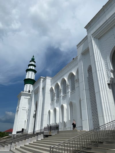 The image shows a mosque with a tall, green-topped minaret on the left, and a white, ornate facade on the right. The mosque is constructed with white stone or concrete and features intricate geometric patterns on its windows and walls. A wide set of concrete stairs leads up to the mosque entrance, with black metal railings on either side. A person wearing a pink hijab and a dark garment is walking up the stairs towards the mosque. The sky above is a mix of blue and white clouds. The location is identified as Aluekambuk, Indonesia.