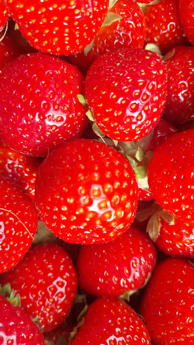 The media file is a close-up, vibrant image of a pile of fresh, ripe strawberries. The strawberries are a deep, glossy red, with their characteristic seeds clearly visible as small yellow dots across their surfaces. Some strawberries have hints of green where their stems or calyxes are attached. The lighting emphasizes the reflective quality of the fruit, creating bright highlights and deep shadows that give the image a sense of depth and texture. The focus is entirely on the strawberries, filling the entire frame and creating an impression of abundance. There is no discernible background, activity, time of day, weather, location cues, emotions, or text. The image is purely a detailed study of the texture and color of fresh strawberries.