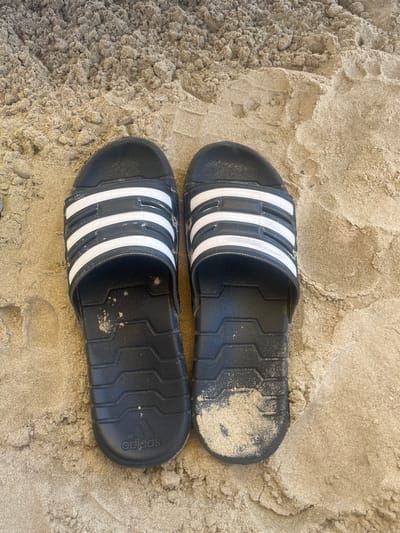A pair of black Adidas sandals with white stripes lie on tan sand. The sandals are positioned next to each other and slightly angled, with one appearing partially covered in sand. The sand is dry and textured. This is likely at the beach in Bashkia Durrës, Albania.