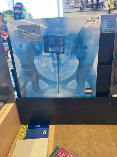 The image depicts the interior of a retail store, likely in the music or entertainment section. The precise location within the city of Edmond, United States, is not discernible from the visual information provided.
The central subject is a vinyl record album displayed upright in a black, multi-tiered retail rack. The album, identified by a sticker as "Lorde - VIRGIN (Bathwater Vinyl)," features a blue-tinted X-ray image of a human pelvis on its cover. A prominent vertical element resembling a zipper extends through the center of the pelvic area, terminating in a T-shaped object consistent with an intrauterine device (IUD) at the bottom. Above the pelvis, a dark, rectangular object resembling a smartphone held by a selfie stick mount is visible. A white price sticker in the top right corner indicates "$44.99" and "NEW," along with a barcode. A black "PARENTAL ADVISORY EXPLICIT CONTENT" label is located in the bottom right corner of the album cover.
To the right of the primary album, a portion of another vinyl record is visible, displaying a tracklist that includes "Dreams," "Linger," "Zombie," "Ode To My Family," and "I Can't Be With You." In the background, out-of-focus plush toys, including green and multicolored characters, are visible on shelves to the left, while other retail products or accessories are stocked on shelving units to the upper right. The foreground shows a light-colored floor, possibly wood or laminate. In the lower-left corner, white dividers with blue and yellow tabs, marked "RAP" and "A," are stacked, indicating music genre or artist categorization. Part of another media case, displaying green and red colors, is visible in the bottom right.