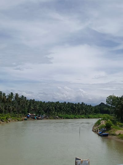 The image depicts a serene river scene in Neuboknaleung, Indonesia, under a cloudy sky. A calm, muddy river flows from the distance towards the viewer, bordered by lush green vegetation on both sides. On the left bank, a dense forest of coconut palm trees stretches across the landscape, with a small cluster of stilt houses and boats docked near the shore. The right bank is also lined with greenery, featuring a rocky embankment and a single blue and white boat moored near the water's edge. In the background, rolling hills are visible, adding depth to the scene. The sky is filled with a mix of white and grey clouds, suggesting an overcast day. There are no visible people in the image. The overall impression is one of tranquility and natural beauty.