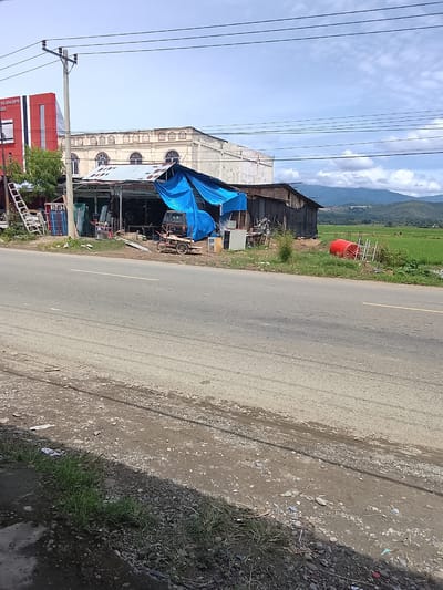 The image depicts a roadside scene in Manyang Cut, Indonesia, on a sunny day with scattered clouds. In the foreground, a two-lane asphalt road shows signs of wear, with a gravelly shoulder on the right and patches of grass and debris.

To the left of the road, a collection of structures and objects are visible. A tall, red and white building with vertical stripes is partially obscured by a tree. Next to it, a small shop or workshop has a corrugated metal roof, partially covered by a blue tarp. Various items are displayed and stored here, including what appear to be window frames, glass panes, and shelving units. A ladder leans against the building. Further down, a small, three-wheeled vehicle with a trailer is parked.

Beyond the workshop, a larger, older building with an arched facade is visible. In the background, a lush green rice paddy stretches out towards rolling hills under a sky with white clouds. The overall scene suggests a semi-urban or rural setting, with commercial activity alongside agricultural land. No people are clearly identifiable in the image.