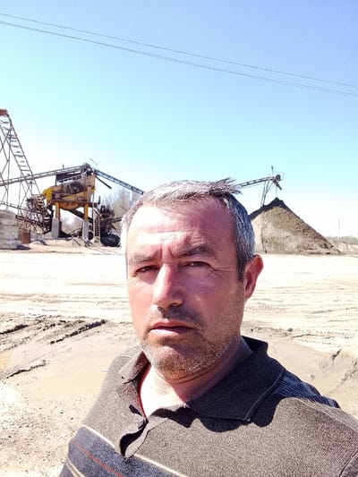 A male individual is depicted in a selfie-style portrait in Yangi Chinoz, Uzbekistan. He is middle-aged with graying hair and a short beard, wearing a dark, striped polo shirt. His gaze is directed towards the camera, and his face is brightly lit. The background presents an industrial aggregate processing facility under clear, sunny skies. Key elements include multiple interconnected conveyor belt structures and processing units, predominantly yellow and dark, along with a large conical pile of light brown aggregate material or earth. The ground consists of light-colored, disturbed soil with visible tracks and some darker, possibly moist, patches. Sparse, bare vegetation is visible behind the machinery. Two thin power lines are visible against the clear blue sky above the industrial structures.