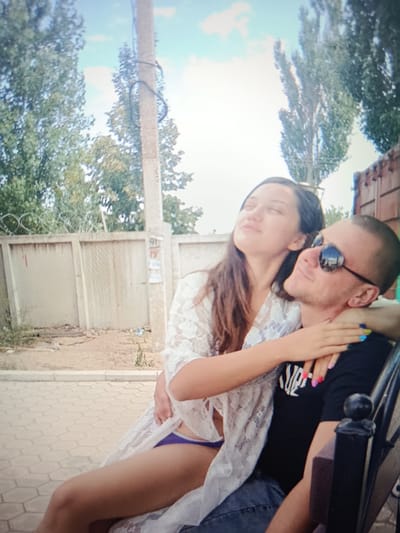 In Pishchane, Ukraine, a couple is seated on a bench outdoors, with a fence and trees in the background. The woman, with long brown hair, is wearing a white lace cover-up over a purple bikini bottom. She has her arm around the man's neck and is leaning her head back, eyes closed. The man, with a shaved head and sunglasses, is wearing a black t-shirt and blue jeans. He has his arm around her waist. They appear to be in a relaxed, intimate moment. The environment suggests a casual, possibly residential or recreational area.