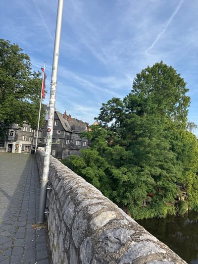 The image shows a view from a stone bridge in Wetzlar, Germany. A large tree with lush green foliage dominates the upper right of the frame, casting shadows on the water below which is reflected in the dark surface. Adjacent to the tree is a row of old, dark-grey buildings with pitched roofs and white-framed windows, typical of German architecture. A lamppost covered in stickers runs vertically through the center of the image, leaning slightly towards the left. Two small flags are attached to the lamppost, one red and one black and red. The sky is a clear blue with wispy white clouds, suggesting it is daytime, possibly late morning or early afternoon. The stonework of the bridge is detailed and weathered, and the path on the bridge is paved with dark grey cobblestones. The scene appears peaceful and serene.