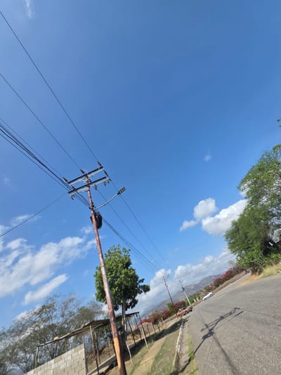 The image depicts a partly cloudy sky over Parroquia Ocumare, Venezuela. A utility pole with attached power lines is visible in the foreground. In the background, a road is lined with greenery. The camera angle suggests an upward view.