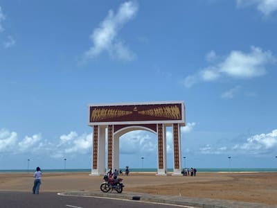 A medium-angle, daytime shot depicts the Gate of No Return, a monument located in Djègbadji, Benin. The monument is a large archway structure, primarily white with dark brown accents and golden figures depicted on its top section. The archway leads to a sandy area with a beach and the ocean visible in the background.
In the foreground, a paved road stretches across the bottom of the frame. To the left of the road, a person in a white shirt and blue jeans stands looking towards the ocean. A motorcycle is parked in front of the monument. Groups of people can be seen standing on the sand beyond the archway. Streetlights are dotted across the landscape. The sky is blue with scattered white clouds.