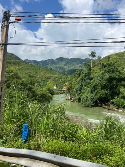The image depicts a rural landscape in Thuận Hòa Commune, Vietnam. A concrete utility pole, adorned with numerous black cables and a small sign bearing the text "CAO QUANG," stands prominently in the left foreground. Beyond the pole, a lush green river flows, bordered by dense vegetation and steep, terraced hillsides covered in trees. A bridge can be seen spanning the river in the mid-ground. In the foreground, a blue plastic-wrapped object, possibly a signpost or marker, is partially visible amidst the tall green grasses. The sky is partly cloudy with white cumulus clouds. A metallic guardrail runs along the bottom of the frame.