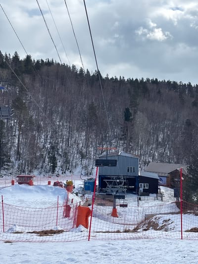 This image depicts a winter ski resort scene in Листвянка, Иркутский region, Russia, on an overcast day. The foreground features snow-covered ground with patches of exposed dry grass and a prominent red safety net on red poles, some with orange padding. In the middle ground, a grey corrugated metal building with a blue base, identified as a ski lift station, stands centrally. Empty ski lift chairs are visible hanging from cables above and near the station. To the right of this building is a smaller, dark-colored wooden building with a gabled roof and a sign displaying a skier. To the left, several red-roofed booths or small structures are positioned on snow mounds. A yellow snow cannon is visible on a snow pile near the central lift station. A single individual in a yellow jacket is partially visible near the snow cannon. Another person in dark attire is obscured near the smaller dark building. The background is dominated by a dense, snow-dusted forest of mixed deciduous (bare) and evergreen trees, extending up a hillside under a cloudy sky. Multiple ski lift cables traverse the upper portion of the frame.