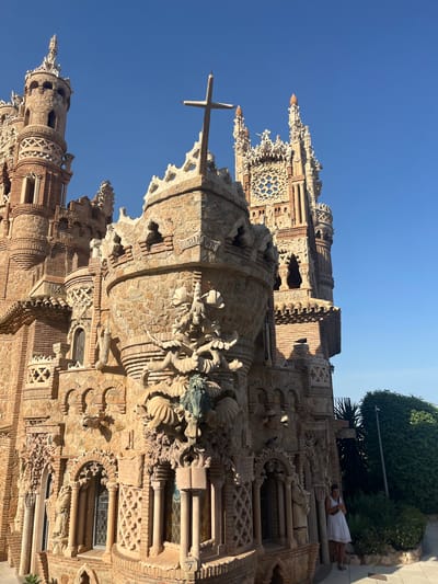 This is a close-up, low-angle photograph of an intricately designed stone and brick building, resembling a castle or a fantastical church. The architecture is characterized by numerous spires, decorative carvings, arches, and latticework windows. A large cross is visible on the top left of the frame, mounted on a decorative pinnacle. The material appears to be a mix of rough-hewn stone and brickwork, giving it a weathered and historic feel.

In the upper right portion of the image, a woman in a white dress is standing, looking towards the building. She is posed on what appears to be a set of stone stairs or a raised platform. Her presence provides a sense of scale to the monumental structure. A pigeon is also visible on the stone facade of the building.

The scene is set against a clear, bright blue sky, suggesting a sunny day. The lighting indicates it is likely mid-day, with strong shadows cast by the architectural elements. Lush green foliage is visible in the background, framing the top right of the building. The overall impression is one of awe-inspiring, unique architecture bathed in natural light. The building's detailed ornamentation suggests it might be a tourist attraction or a significant cultural site. The location cues and architectural style are consistent with Benalmádena, Spain, a region known for its distinctive landmarks. No visible text is legible in the image.