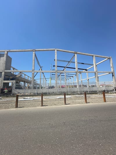 The image shows the skeletal framework of a large building under construction in the city of Poti, Georgia. The structure is primarily composed of concrete beams and columns, with some steel beams also visible. A crane is present at the site, indicating ongoing construction activity. A fence with a decorative, circular design separates the construction area from a road in the foreground. The sky is clear and blue, suggesting a sunny day.