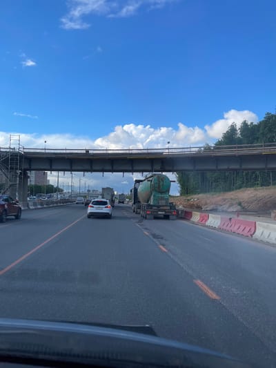 The image shows a view from inside a vehicle traveling on a road, likely in Novodrozhzhino, Russia given the prompt. Above the road, a bridge is visible, possibly under construction, with scaffolding present. The sky is partially obscured by the bridge, revealing blue sky and white clouds. Traffic is present on the road, including a white car directly ahead and a green truck further down the road. A row of red and white road barriers are also along the side of the road. Buildings can be seen in the distance.