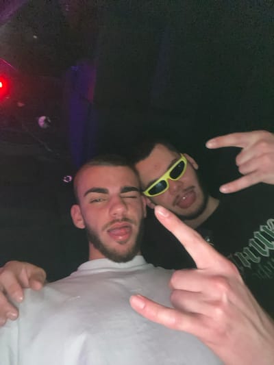 The image displays two young adult males posing in a dimly lit indoor environment, consistent with a nightclub or entertainment venue in Пампорово, Bulgaria. The male on the left has light skin, short dark hair, and a beard. He wears a light-colored t-shirt, winks with his right eye, and extends his tongue from his mouth. His left arm is around the shoulder of the male on the right. The male on the right is positioned slightly behind and to the right, wearing narrow neon yellow sunglasses and a dark t-shirt with a partially visible green graphic. He also extends his tongue and forms a "rock on" hand gesture with his right hand. The background features ambient red and purple lighting, indicating low-light conditions.
