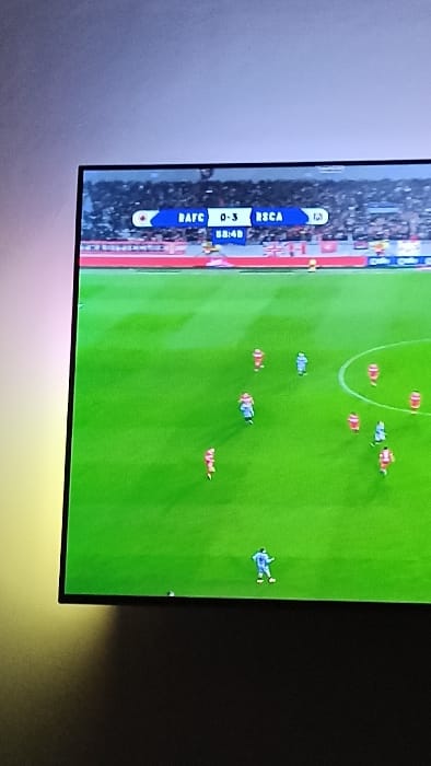 A television displays a soccer match, showing an aerial view of a green field with players in red and blue uniforms. The score is RAFC 0-3 RSCA, with the game time at 88:48. The screen is mounted on a wall with ambient lighting emitting from behind the television.
