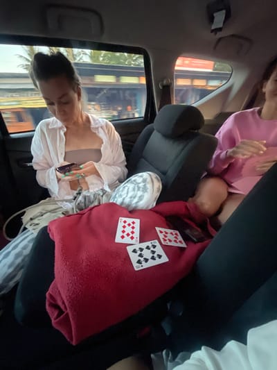 Two women are seated in the back of a vehicle, which appears to be moving, judging by the blurred scenery outside the windows, suggesting it is a roadway in Pekutatan, Indonesia. The woman on the left is looking down at her phone, wearing a light-colored open shirt over a tan top and striped pants. She has green nail polish. In front of her, on a red blanket, are three playing cards: a 10 of diamonds, a 10 of spades, and another 10 of spades. There is also a black object, possibly a phone, on the blanket. The woman on the right is wearing a pink sweatshirt and is partially visible, looking towards the front of the vehicle and smiling. The interior of the vehicle is dark, with visible seat upholstery and headrests. The windows show palm trees and a building with red and blue elements, consistent with the tropical environment of Pekutatan.