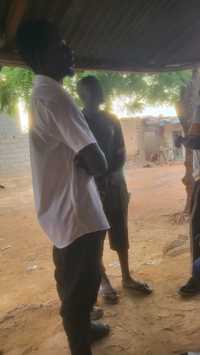 The image shows at least two people standing outside in Jogana, Nigeria. One person is wearing a white shirt and black pants, and another person is wearing a green shirt and brown pants. There are also people wearing sandals, one of them also wearing shoes. The ground is dirt. In the background, there are buildings. The photo seems to be taken from under a corrugated metal roof.
